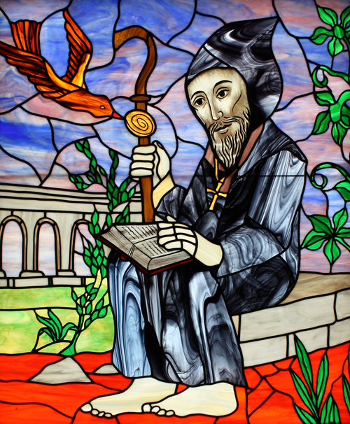 Image of a stained glass window showing a stylized Benedict in his abbot's habit, tended to by a raven.