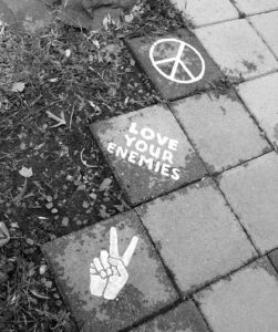 Chalk on sidewalk tiles -- a peace sign, fingers making the peace sign, and the words "Love your enemies"